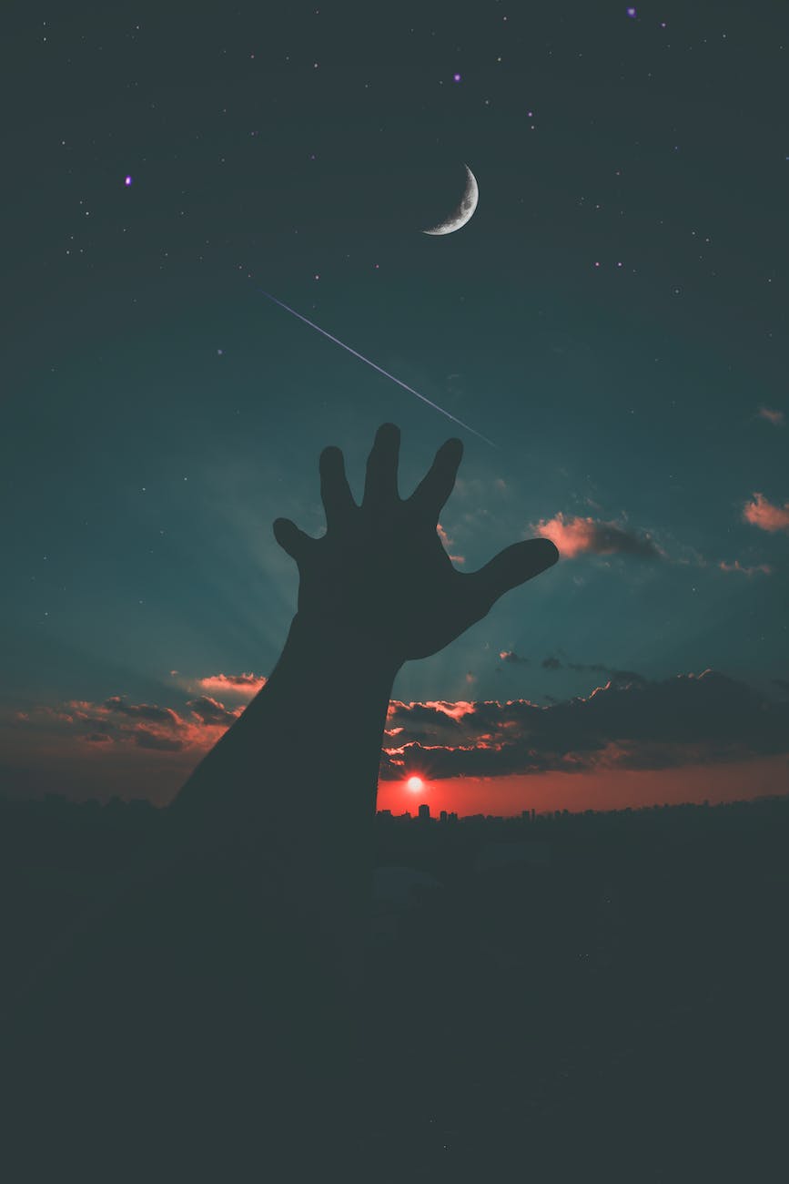 A silhouetted hand reaching towards a crescent moon against a twilight sky filled with stars and wispy clouds, with a setting sun casting an orange glow on the horizon.