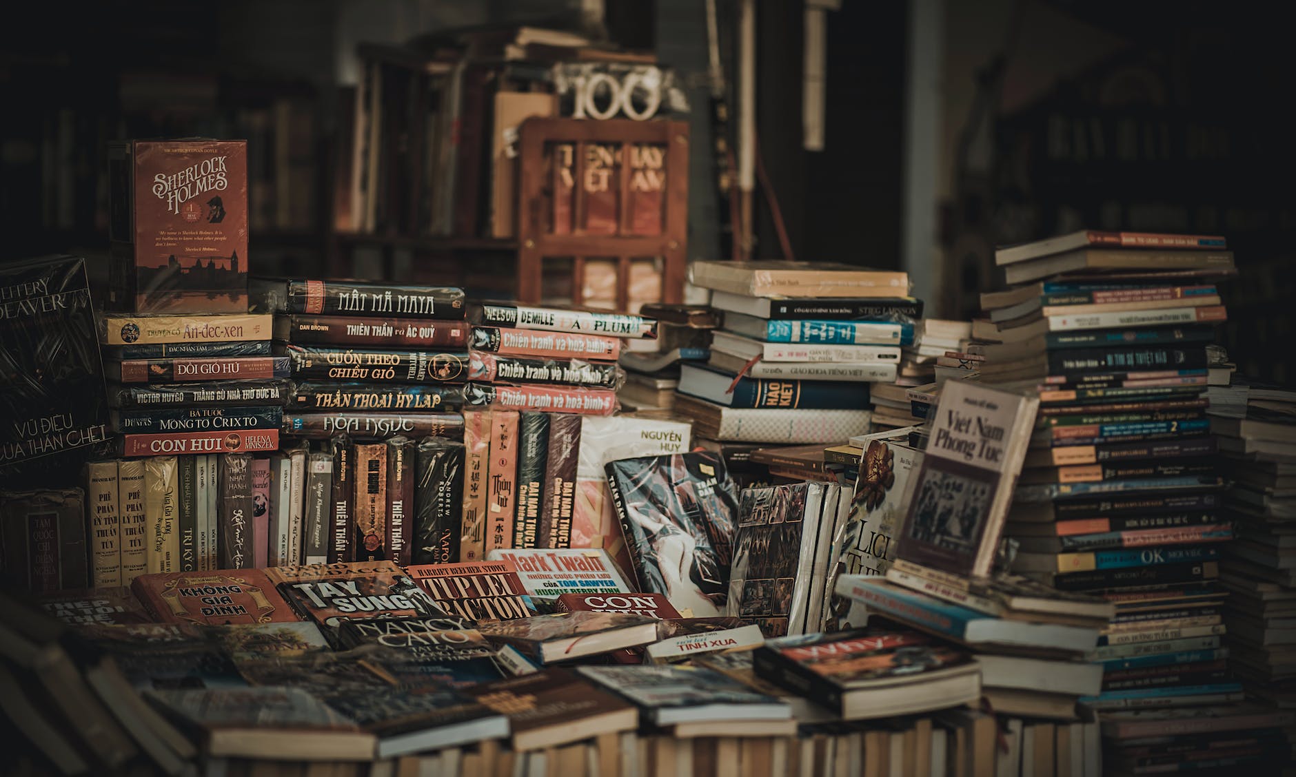 A cluttered collection of books stacked in various positions, with some titles visible including 'Sherlock Holmes' and other literary works, set in a dimly lit interior.